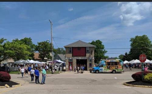 A bustling outdoor market scene with tents, people, and a colorful food truck near a red building on a sunny day.
