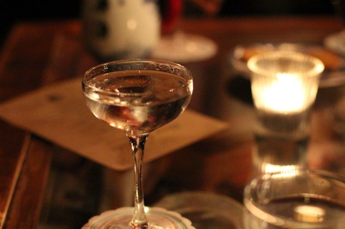 A close-up of a cocktail glass filled with a clear drink, surrounded by soft candlelight and other glassware.