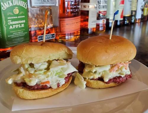 Two sliders with coleslaw on soft buns, served on a plate with a bar backdrop featuring various liquor bottles.