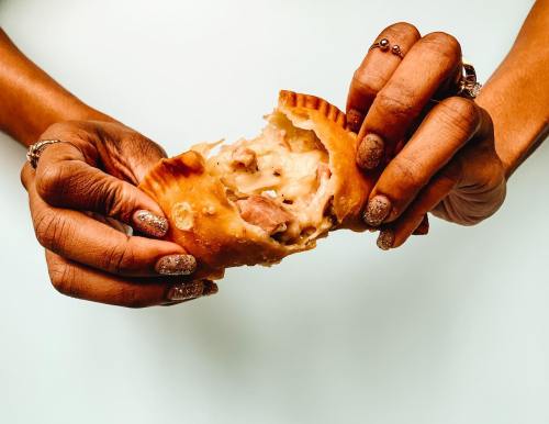 A person holds a partially torn empanada, revealing a creamy filling, with manicured nails and rings on their fingers.