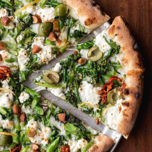 A close-up of a pizza topped with ricotta, green vegetables, olives, and almonds on a wooden surface.