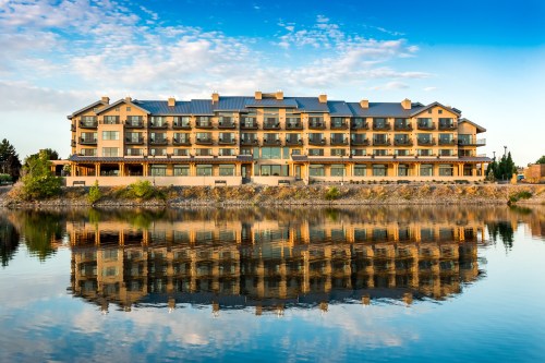 A modern hotel by a calm lake, reflecting its image on the water under a blue sky with scattered clouds.