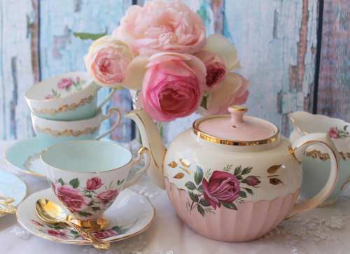 A vintage tea set with floral designs, including a teapot, cups, and a bouquet of pink roses on a rustic background.