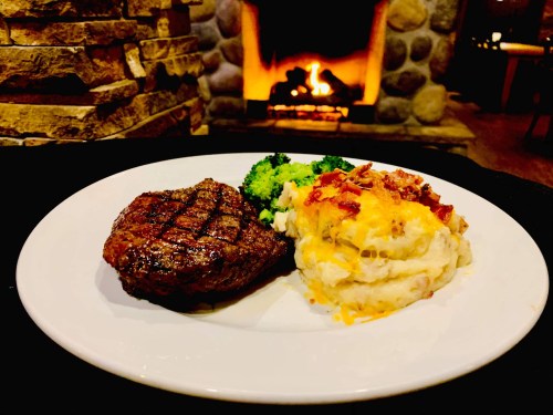 A plate with a grilled steak, creamy mashed potatoes topped with cheese, and steamed broccoli, with a fireplace in the background.