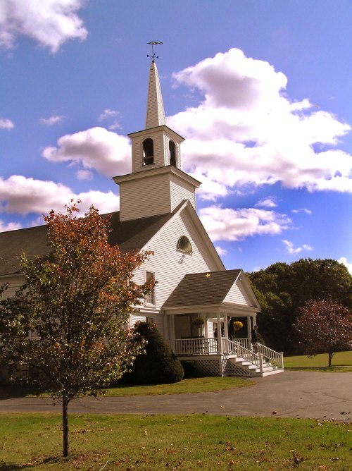 A white church with a tall steeple, surrounded by trees and a blue sky with fluffy clouds.