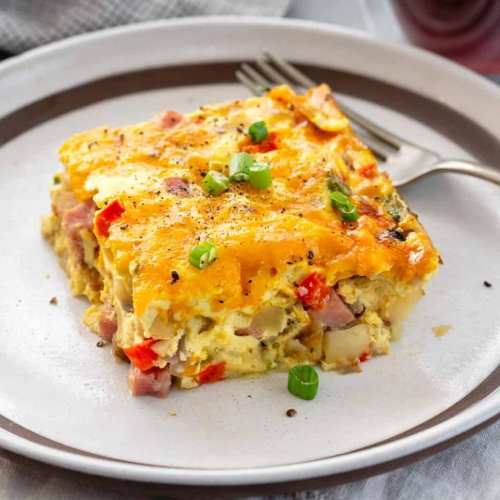 A slice of cheesy ham and vegetable casserole topped with green onions on a plate, with a fork beside it.