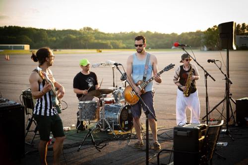 A band performs outdoors at sunset, featuring a guitarist, drummer, and saxophonist on an empty airstrip.