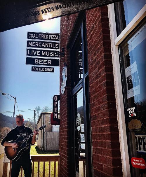 A musician plays guitar outside a restaurant with signs for pizza, live music, and beer. Bright sunny day.