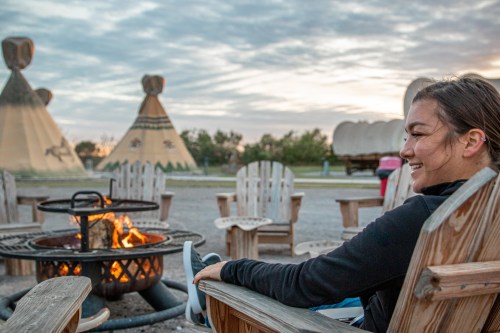 A person sits in an outdoor chair by a fire pit, with teepees in the background under a cloudy sky.