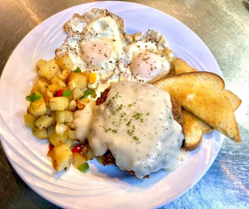 A plate of breakfast featuring two fried eggs, country gravy over a biscuit, and diced potatoes with toast.