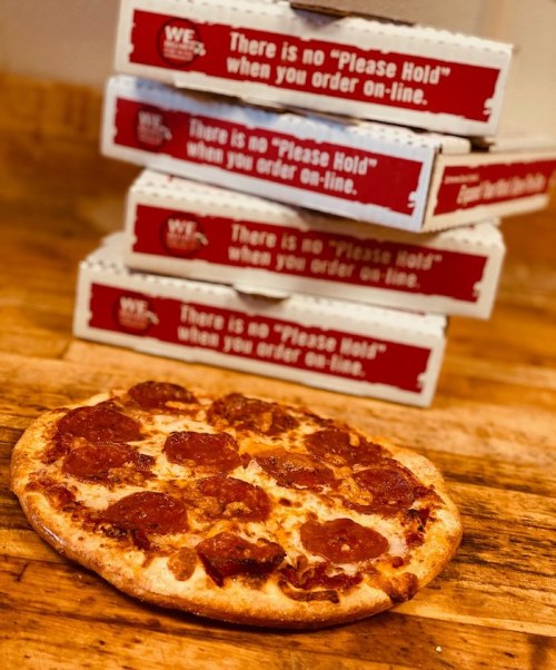 A pepperoni pizza on a wooden surface with stacked pizza boxes in the background, featuring a humorous message.