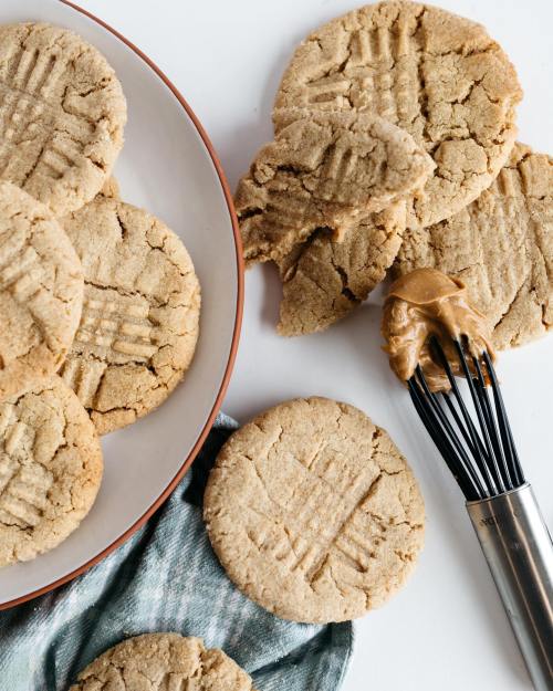 A plate of freshly baked peanut butter cookies with a whisk and a dollop of peanut butter on a cloth.