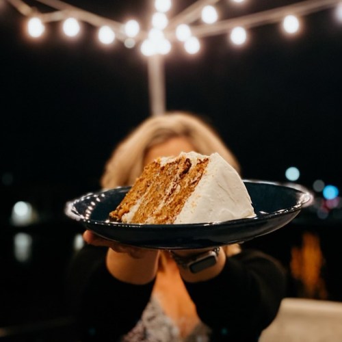 A person holds a plate with a slice of cake, illuminated by string lights in the background at night.