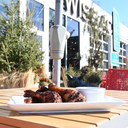 A plate of grilled chicken drumsticks on a table outside, with greenery and a restaurant in the background.