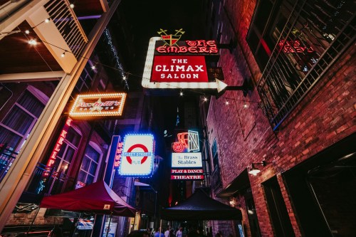 Colorful neon signs illuminate a narrow alleyway, featuring "The Climax Saloon" and "The Brass Stables."