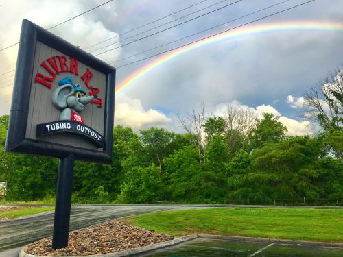A colorful sign for "River Rat Tubing Outpost" with a rainbow in the background and lush greenery surrounding it.