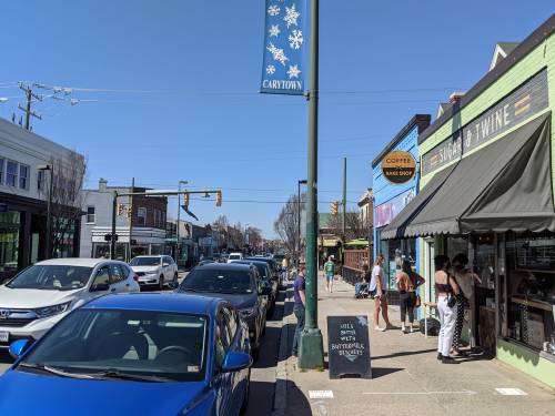 A busy street scene with cars parked along the curb, shops, and people enjoying a sunny day.