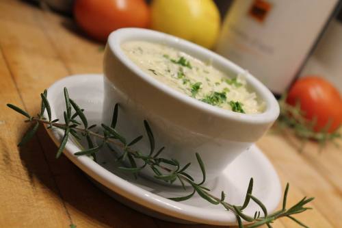 A small bowl of creamy dip garnished with herbs, placed on a white plate, with fresh tomatoes and lemon in the background.