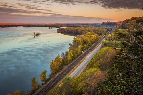 A scenic view of a river surrounded by trees, with a railway and road running alongside, under a colorful sunset sky.