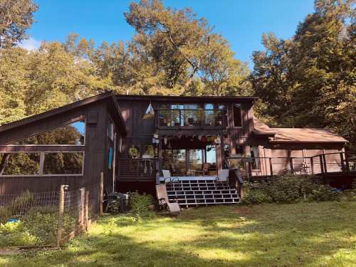 A modern wooden house surrounded by trees, featuring a deck and large windows, set in a lush green landscape.