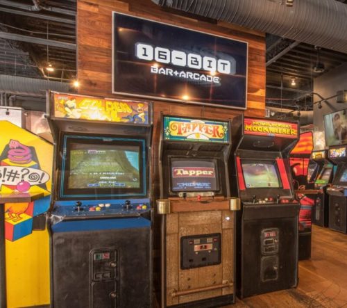 A row of vintage arcade machines in a bar setting, featuring games like "Golden Axe" and "Tapper."