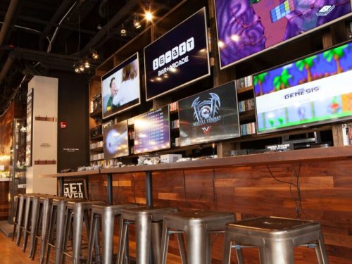 A modern arcade with multiple screens displaying games, and metal stools lined up at a wooden counter.