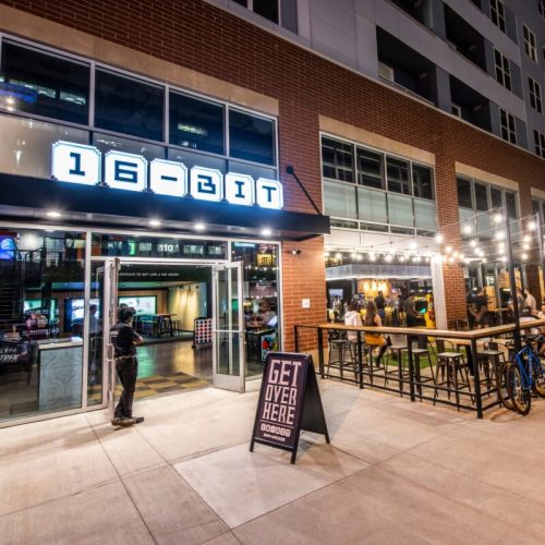 Exterior of 16-Bit bar with neon sign, outdoor seating, and a sign reading "GET OVER HERE" in a lively urban setting.