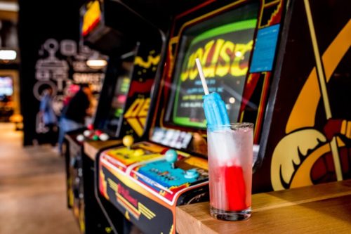A colorful drink with a popsicle sits on a table in front of vintage arcade machines.