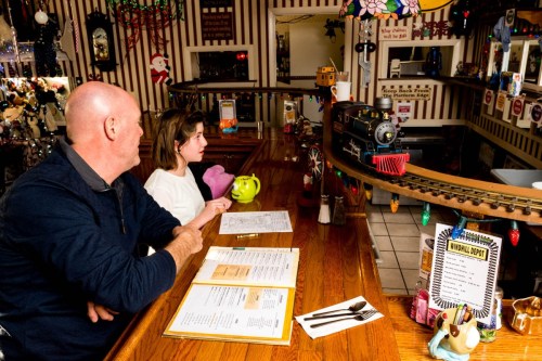 A man and a girl sit at a bar, watching a toy train on a track above them, with festive decorations around.