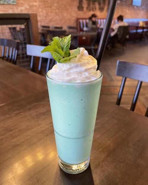 A tall glass of mint green shake topped with whipped cream and a mint leaf, set on a wooden table in a cozy café.