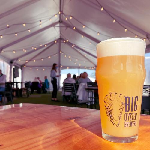A glass of beer sits on a wooden table, with a tented event space and people in the background.