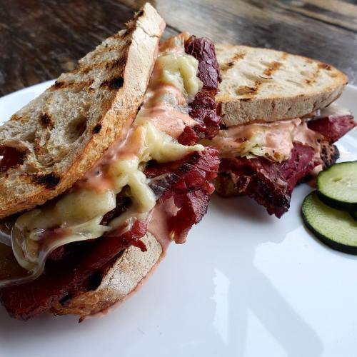A close-up of a grilled sandwich with layers of meat, cheese, and sauce, served with pickles on the side.