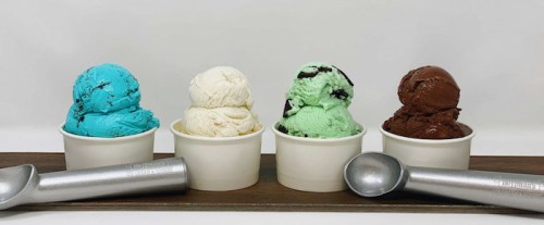 Four cups of ice cream in blue, white, green, and brown, with metal scoops beside them on a wooden tray.