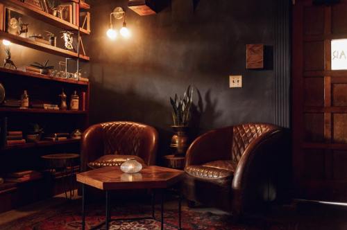 Cozy interior with two leather chairs, a wooden table, and shelves filled with decor against a dark wall.
