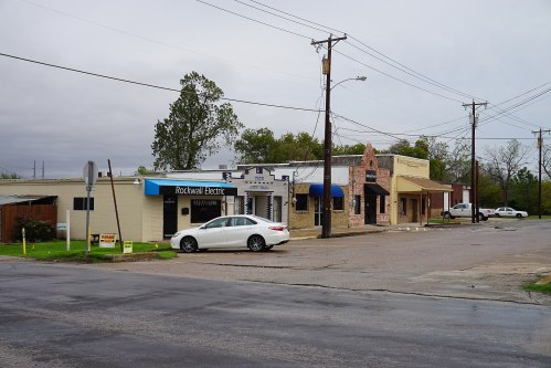 A small town street featuring several businesses, including Rockwall Electric, under a cloudy sky.
