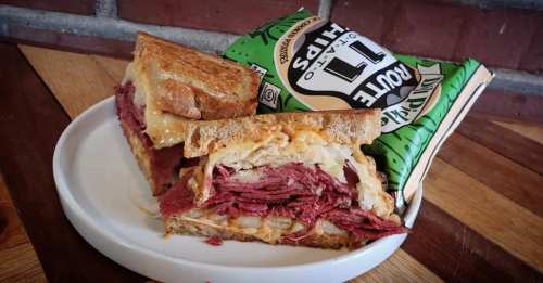 A hearty sandwich with layers of corned beef, sauerkraut, and melted cheese, served with a side of potato chips.
