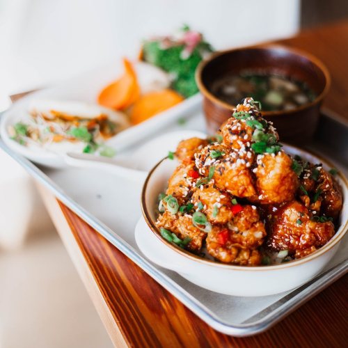 A tray with a bowl of sesame chicken, side dishes of vegetables, and a small bowl of soup.