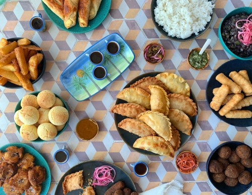 A vibrant spread of various dishes, including pastries, rice, and sauces, arranged on a patterned table.