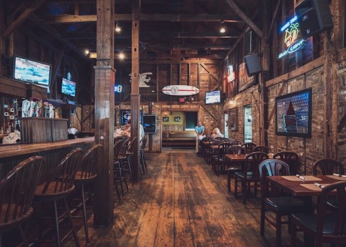 A rustic restaurant interior with wooden beams, a bar, and neon signs, featuring empty tables and a surfboard on the wall.