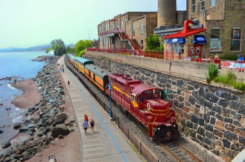 A vintage train travels along a waterfront, with people walking on a path beside the rocky shore and buildings in the background.