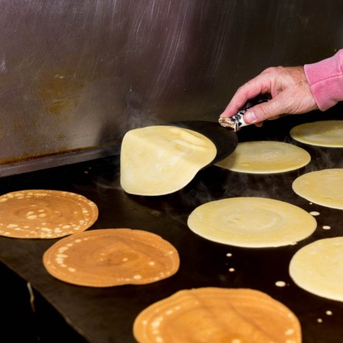 A hand using a spatula to flip pancakes on a griddle, with various pancakes cooking in the background.