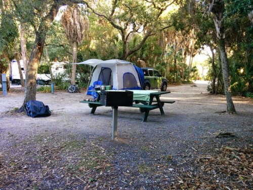 A campsite with a tent, picnic table, and grill surrounded by trees and a gravel path.
