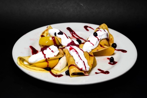 A plate of crepes filled with fruit, topped with whipped cream and berry sauce, against a dark background.