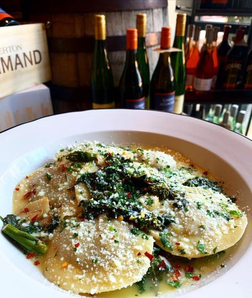 A bowl of ravioli topped with herbs, cheese, and spices, with wine bottles in the background.