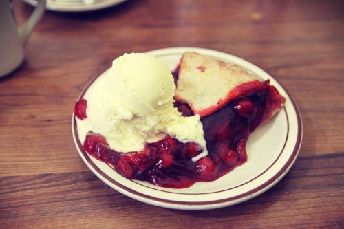 A slice of cherry pie topped with a scoop of vanilla ice cream on a plate, set on a wooden table.