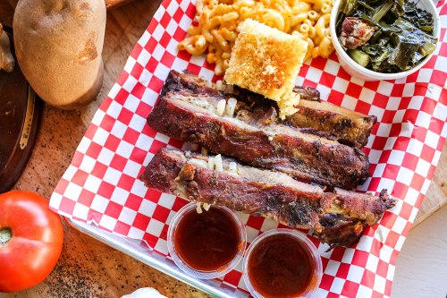 A plate of barbecue ribs with macaroni and cheese, cornbread, collard greens, and two cups of sauce on a checkered tray.