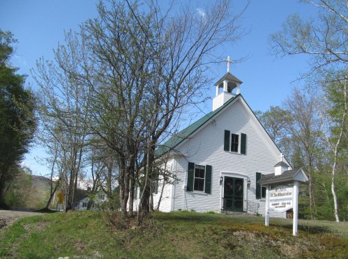A white church with a green roof and steeple, surrounded by trees and a sign in front, set in a rural landscape.