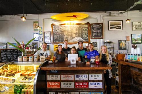 A group of six people stands behind a café counter, smiling, with a menu board and surf-themed decor in the background.
