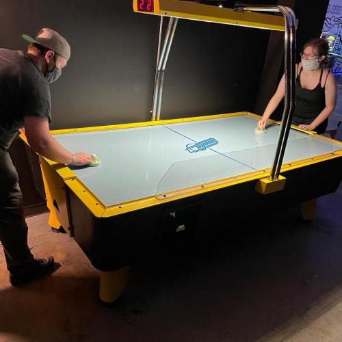 Two people play air hockey at a table, wearing masks, with a bright yellow and black design in a dimly lit room.