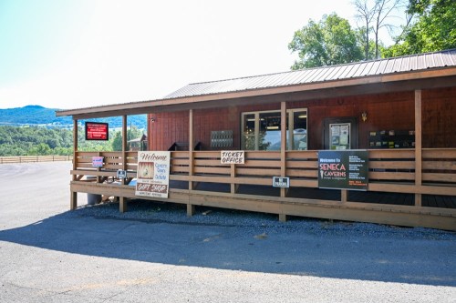 A wooden ticket office with signs, surrounded by trees and mountains in the background on a sunny day.